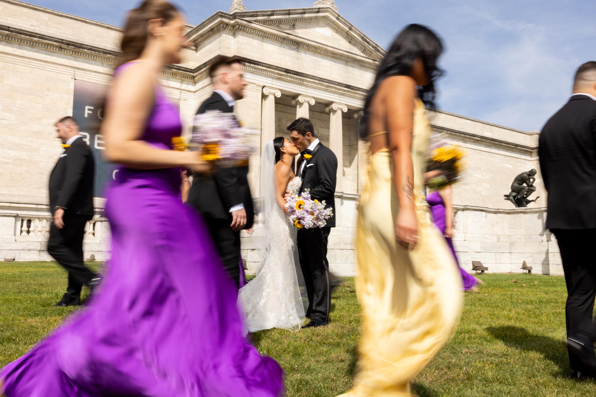 A wedding party crossing the museum lawn mid-stride while the couple kisses